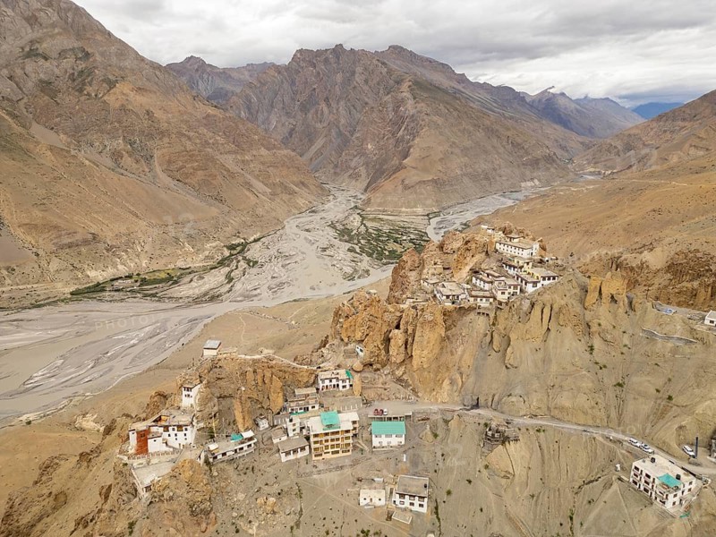 A stunning aerial view of Dhankar village and monastery nestled atop rocky cliffs in Spiti Valley, India. The rugged landscape and meandering river create a picturesque scene