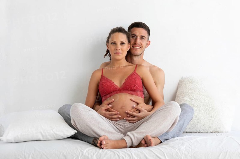 A couple sits on a bed, the pregnant woman in comfortable loungewear, lovingly embraced by her partner A serene moment of togetherness and anticipation unfolds in a bright room