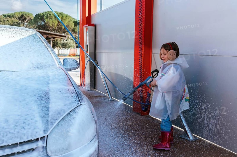 A young child wearing a raincoat and earmuffs joyfully sprays a car with a high pressure hose at a car wash. The car is covered in soap, creating a bubbly scene