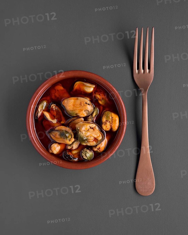 Top view of a bowl filled with mussels covered in a vibrant sauce, accompanied by a wooden fork on a dark gray background