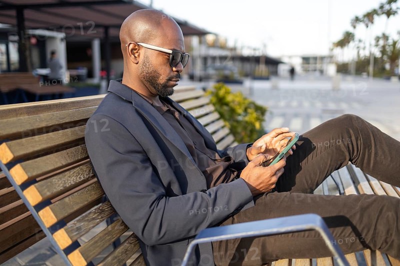 A cuban man wearing sunglasses and a suit relaxes on a park bench, attentively looking at their smartphone. The casual outdoor setting suggests a break from work