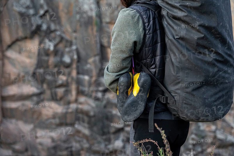 A climber prepares for a traditional climbing adventure in nature, carrying essential gear including climbing shoes and a large backpack, set against a rugged rock face