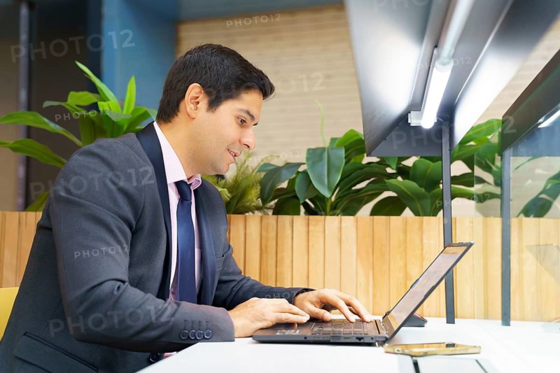 A young Chilean businessman in a modern suit focuses on his laptop in a contemporary setting, surrounded by plants, embodying professionalism and concentration