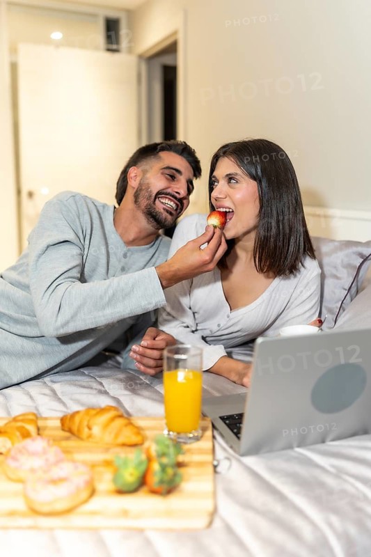 Hispanic man giving a strawberry to his smiling girlfriend while having breakfast in bed and using a laptop