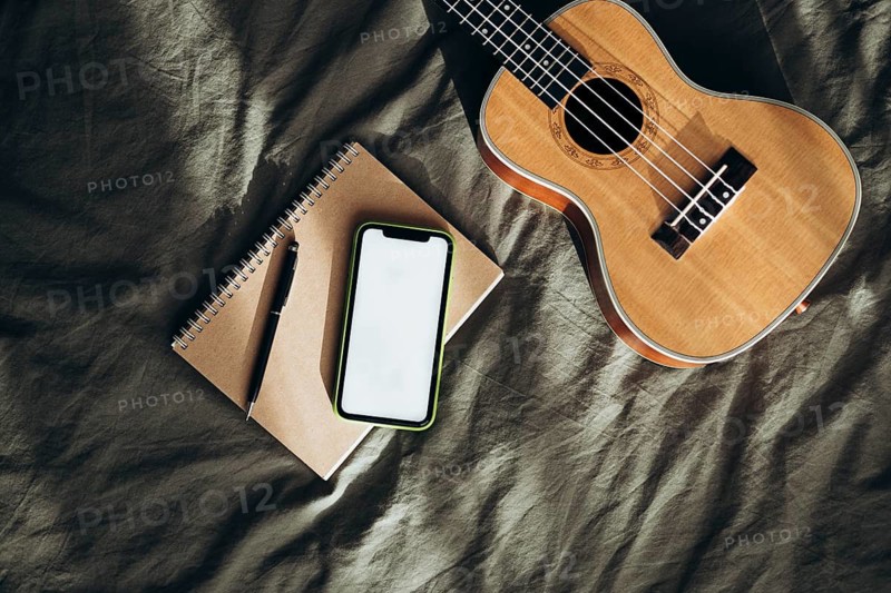 A wooden ukulele, notebook with a pen, and a smartphone lie on a textured fabric surface The arrangement captures creativity, inspiration, and everyday moments