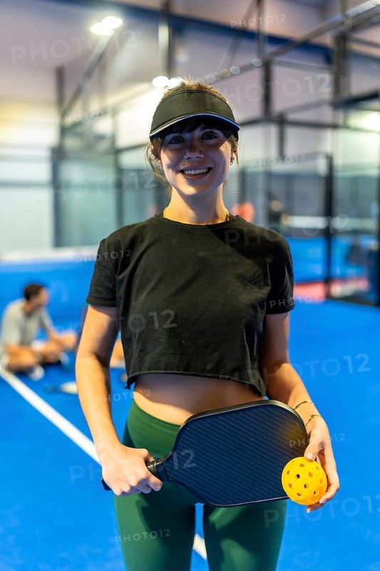 Young woman standing on an indoor blue pickleball court, smiling while holding a paddle and a ball, ready to engage in a dynamic and active sporting game