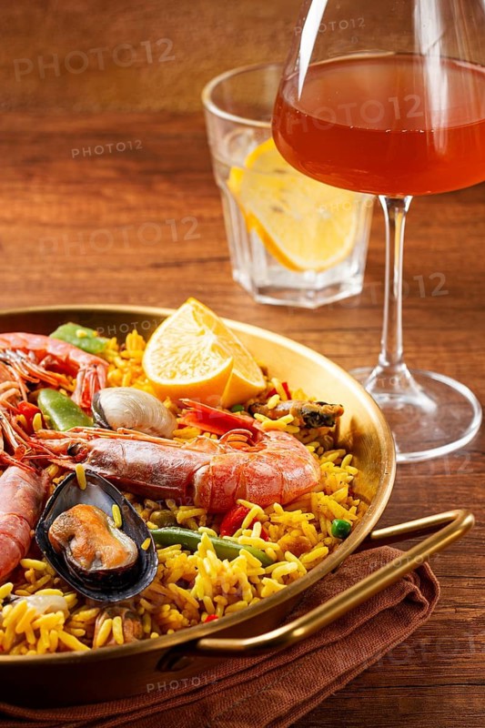 A large dish of seafood paella features shrimp, mussels, and other ingredients. A glass with a drink and slice of lemon is nearby on the wooden table. The meal is ready to eat