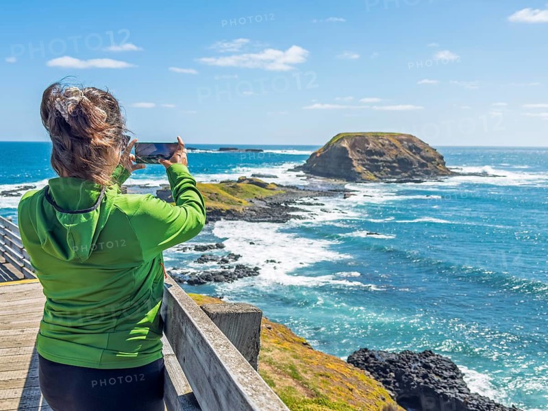 A woman in a green jacket photographs a scenic coastal view with her smartphone in Phillip Island, Australia. The setting features clear blue skies, ocean waves, and rugged cliffs