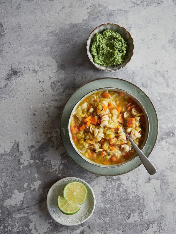 Plate of delicious vegetable and pasta soup called minestrone served on a grey countertop with green pesto sauce and lime slices, top view