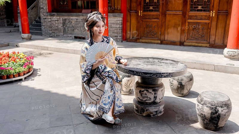 A woman dressed in Qing Dynasty attire is seated outdoors, holding a decorative fan in Pingyao, China. She is surrounded by traditional architecture and vibrant flowers