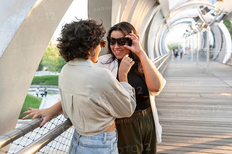 A joyful lesbian couple shares a playful moment on a modern, arched bridge. Their connection is evident in their smiles, set against a sunny backdrop with architectural details