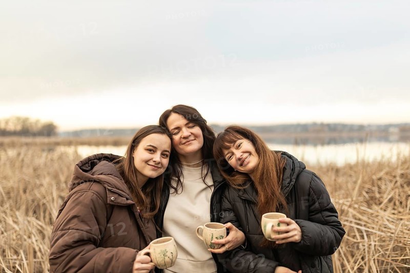 Three female friends enjoy a cozy gathering outdoors, holding mugs and sharing smiles against a scenic lake background. They appear content and relaxed, embodying a peaceful moment together