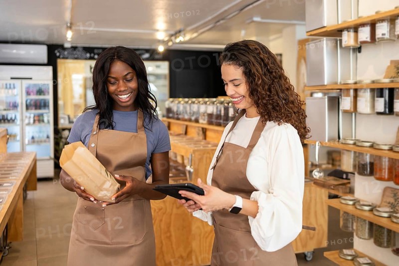 Two friendly clerks wearing aprons stand inside an organic bulk store. One holds a tablet while the other holds a paper bag, smiling as they discuss their work