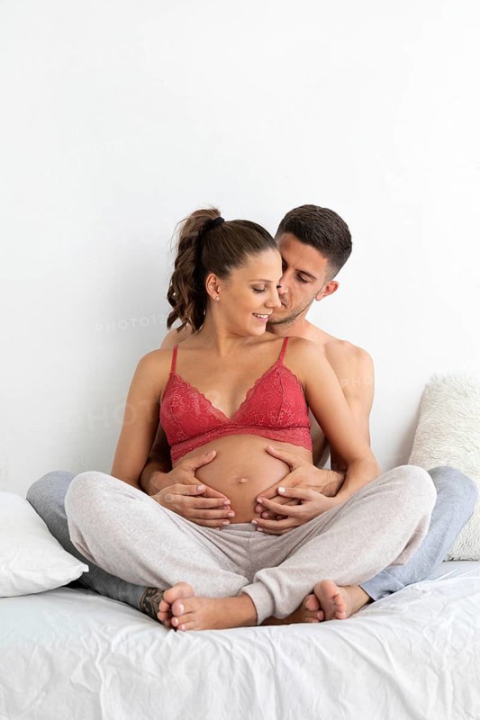 A tender moment shared by a couple, with the man embracing the woman from behind, highlighting her pregnant belly They sit comfortably on a bed, symbolizing love and anticipation