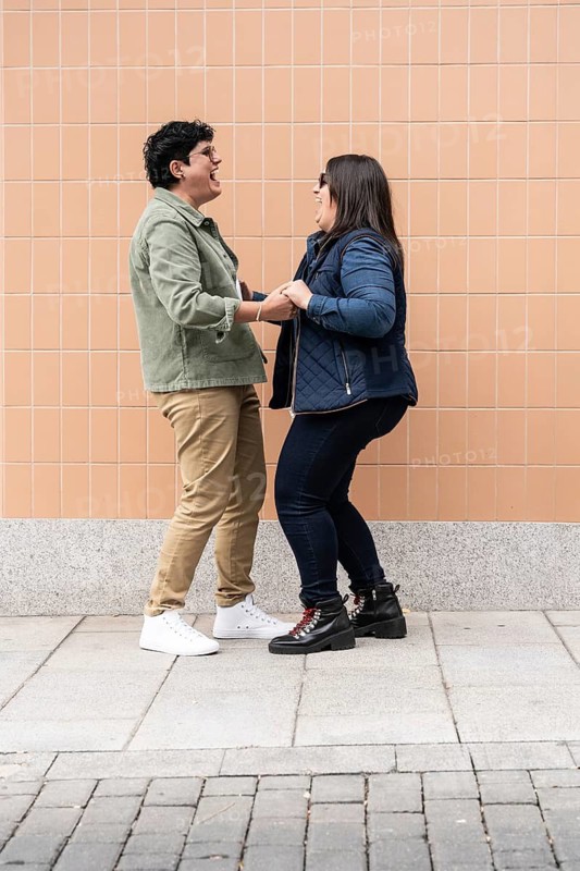 A joyful moment between a lesbian curvy couple, holding hands and laughing warmly against a tiled wall background, capturing the essence of love and happiness
