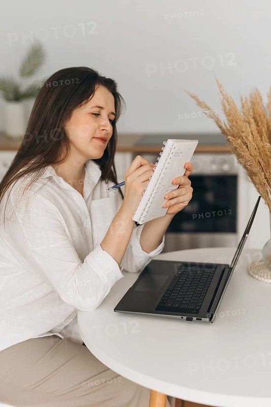A nutritionist sitting at a table, writing in a notebook while using a laptop. The setting is a bright kitchen, emphasizing a professional yet cozy atmosphere
