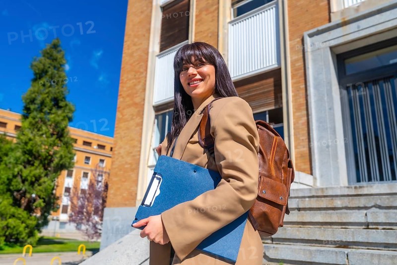 Young woman smiling while walking up steps on university campus, wearing a jacket and carrying a folder along with a brown backpack, representing education and academic life