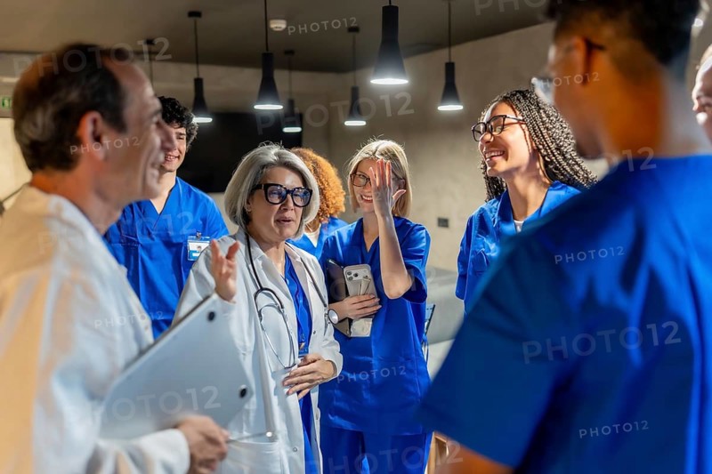 Group of multi ethnic healthcare professionals, including doctors, nurses, and medical students, engaging in a lively discussion inside a modern hospital or clinic environment
