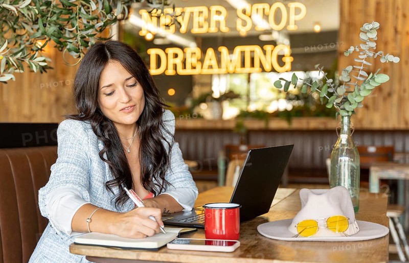 A woman in a cozy cafe, writing in a notebook with a laptop open, a red mug beside her. Behind her, a neon sign reads Never Stop Dreaming, creating a warm, motivational ambiance