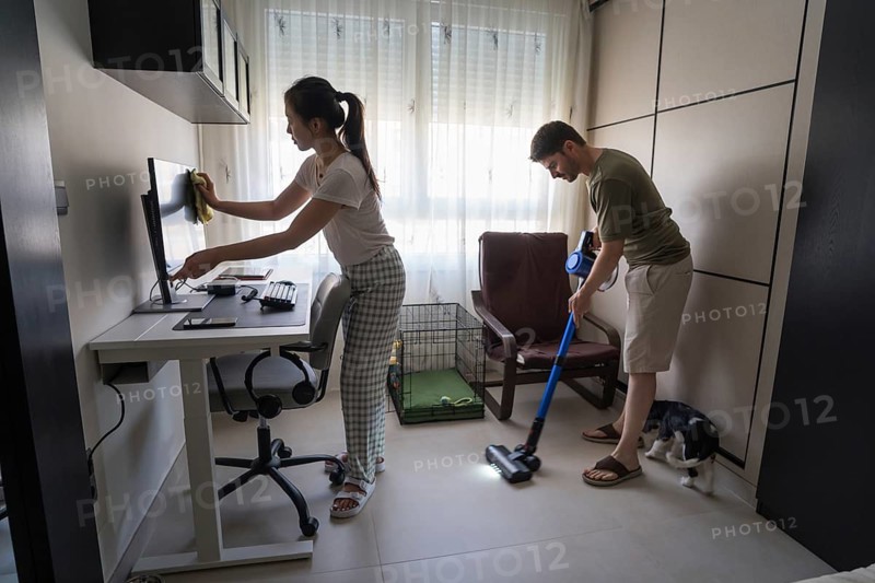 A modern couple engaged in household chores, showcasing shared responsibilities. One partner vacuums while the other cleans a computer, illustrating teamwork in house cleaning