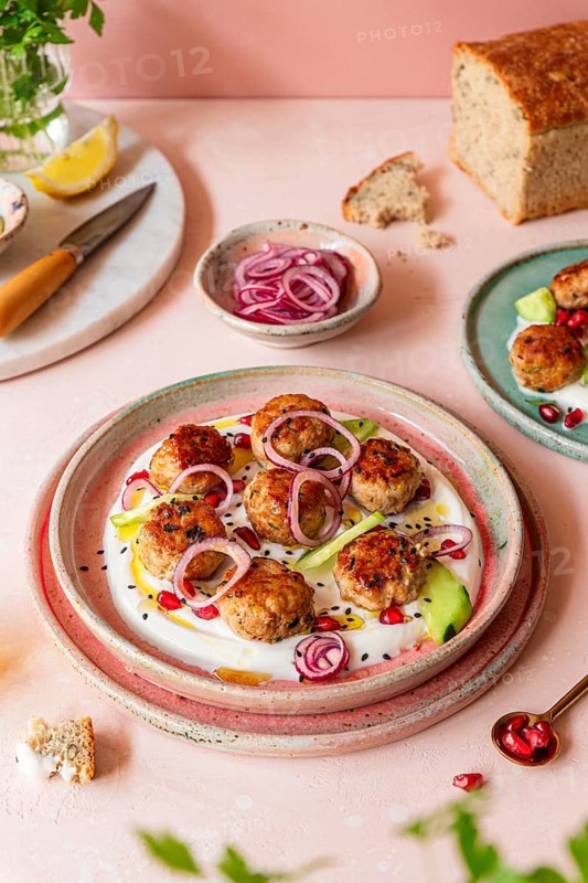 Savory meatballs on a creamy yogurt sauce, topped with sliced red onions, pomegranate seeds, and cucumber, creating a vibrant culinary presentation on a ceramic plate