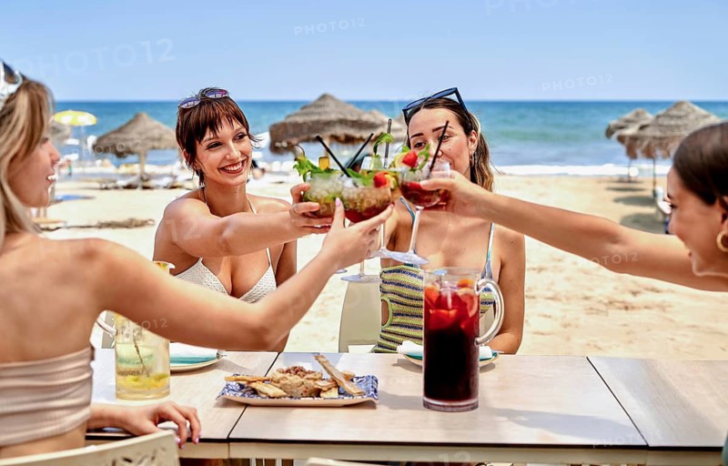 Group of friends enjoy a lively gathering on the beach, raising their vibrant cocktails and savoring delicious snacks while soaking up the sun and sea breeze