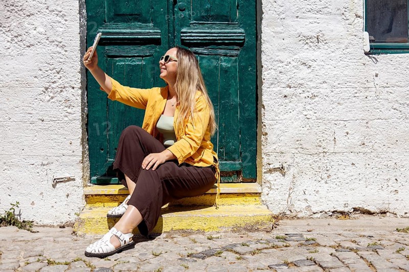 A cheerful woman tourist sits on cobblestone steps, taking a selfie in front of a rustic green door, enjoying a sunny day. Her casual outfit reflects a carefree spirit