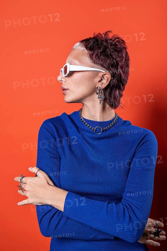 A fashionable young woman in a vivid blue top stands against an orange background, showcasing her unique earrings, necklace, and rings. Her bold, modern haircut and white sunglasses add flair