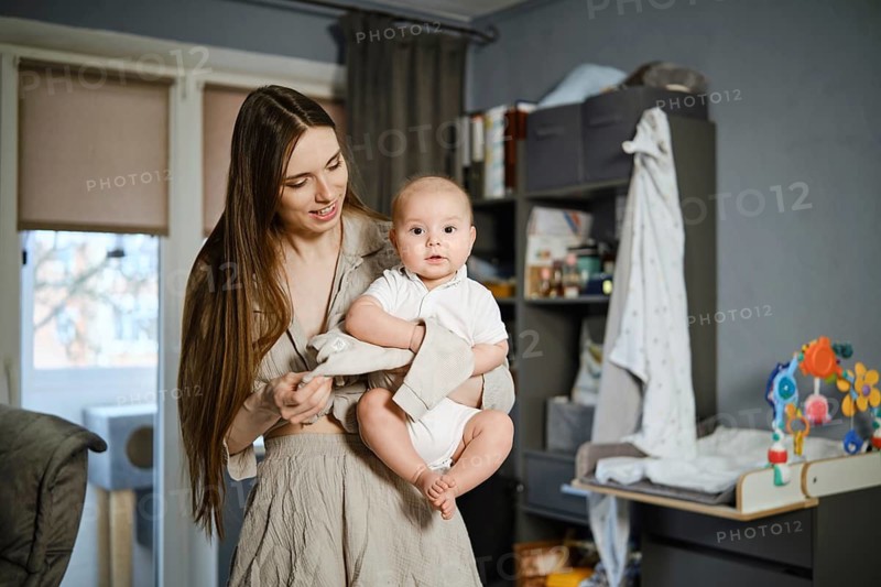 A mother with long hair stands in a living room holding her baby. The baby wears a white outfit and looks around. The room has shelves and curtains