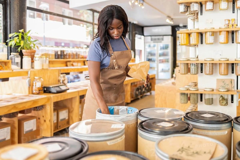 A female store clerk wearing an apron is organizing items in an inviting organic bulk store, surrounded by large containers and shelving filled with various natural products