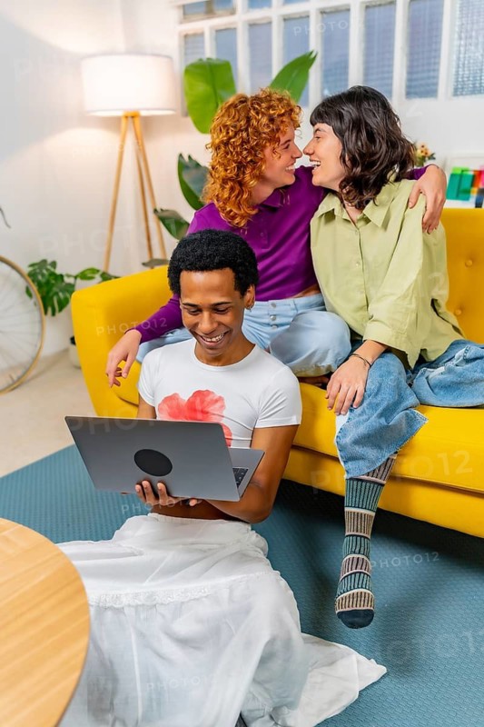 Vertical photo of a Lesbian couple about to kiss next to transgender friend using laptop at home
