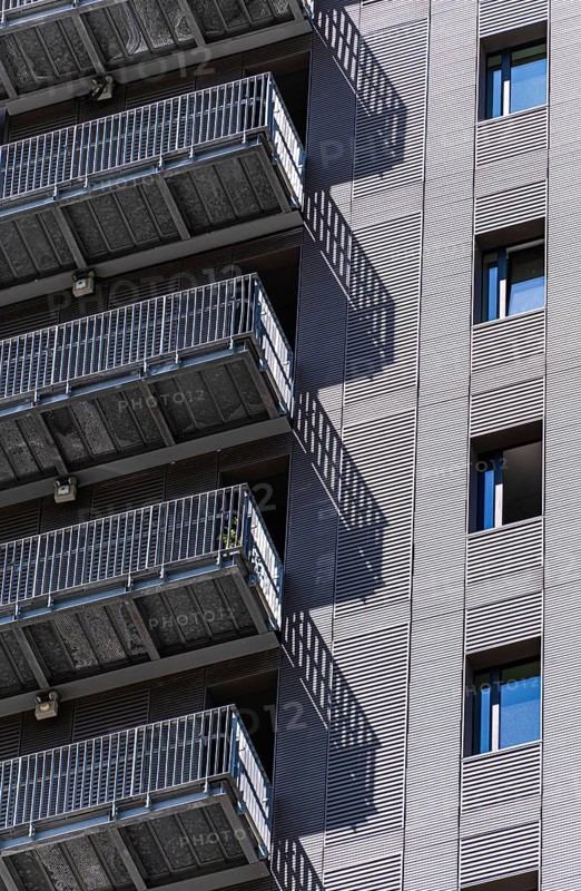 Close-up of a modern urban building facade with metal stairs casting shadows on the wall. The image highlights the architectural design and texture of the metal elements