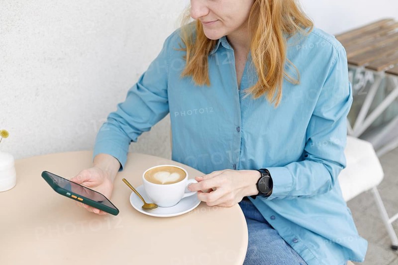 A woman in a blue shirt sits at a cafe table in Warsaw, holding a smartphone and a coffee cup adorned with latte art A smartwatch is visible on her wrist