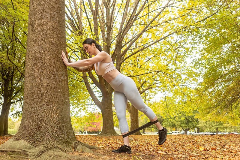A woman exercises using a resistance band in an autumn park in New Zealand. Surrounded by vibrant fall foliage, she enjoys a refreshing outdoor workout amidst nature beauty