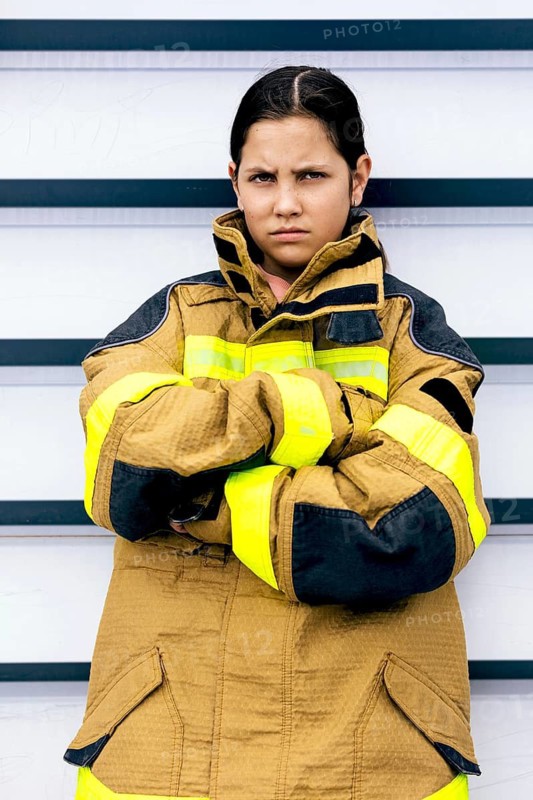 A young girl in a firefighter suit stands confidently with crossed arms. Her serious expression and professional attire convey determination and readiness for challenges
