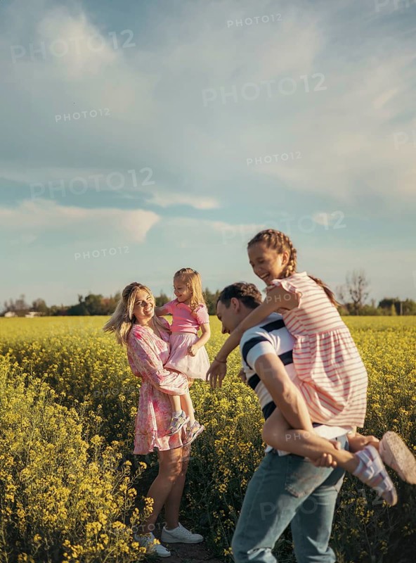 A happy family plays together in a beautiful yellow flower field. The father carries his daughter on his back while the mother holds the youngest, capturing love, fun, and connection