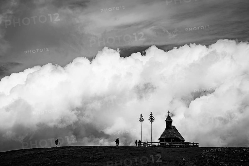 A dramatic black and white landscape featuring a small wooden chapel on a ridge, silhouetted against massive, towering white clouds with small figures of hikers nearby in Slovenia