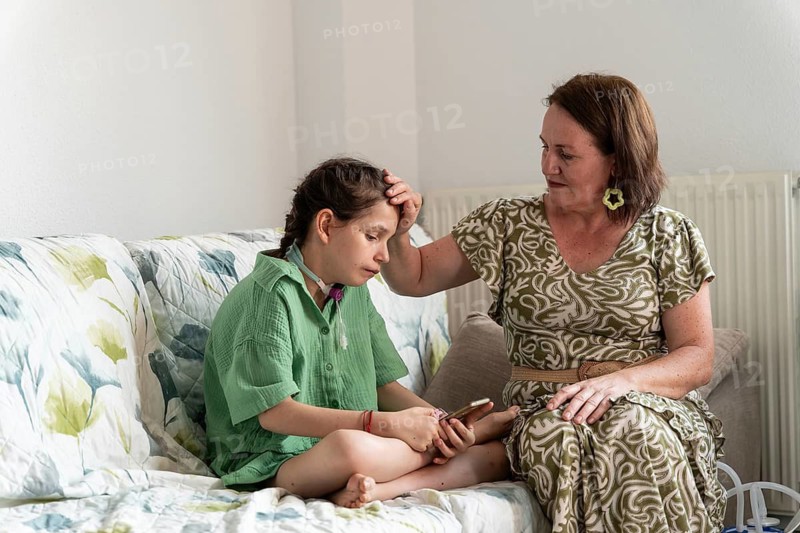 A caring family scene featuring a girl with a tracheostomy sitting beside a supportive mother. The atmosphere is filled with warmth and empathy