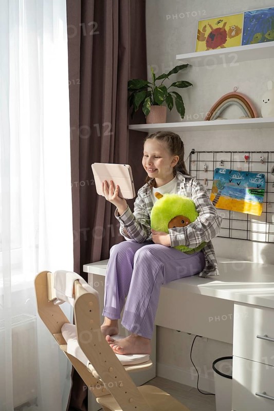 A young girl sits at home, happily engaging with a tablet while hugging a plush toy. Sunlight streams through a window, creating a warm, cozy atmosphere