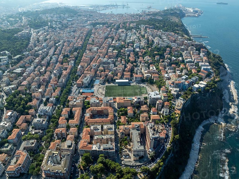 A stunning aerial view of Ancona, showcasing its vibrant architecture, coastline, and lush greenery. The city stretches along the Adriatic Sea, with clear skies above