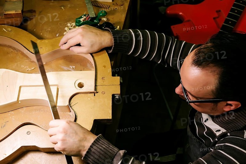A luthier meticulously shapes a bass guitar body, showcasing the art of craftsmanship. The workshop table holds tools and woodworking materials, embodying dedication to detail