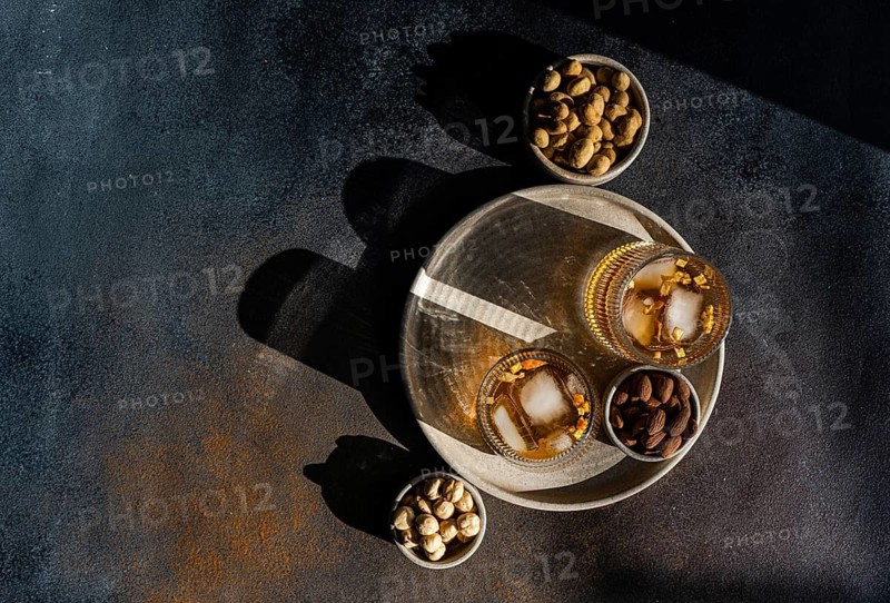Top view of spiced cognac based cocktails with cinnamon and star anise, accompanied by bowls of almonds, peanuts, and hazelnuts, served on a rustic tray set against a dark textured background