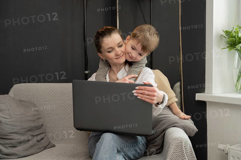 A mother is sitting on a couch using a laptop, while her young child affectionately hugs her from behind. The scene captures a perfect blend of work and family time at home