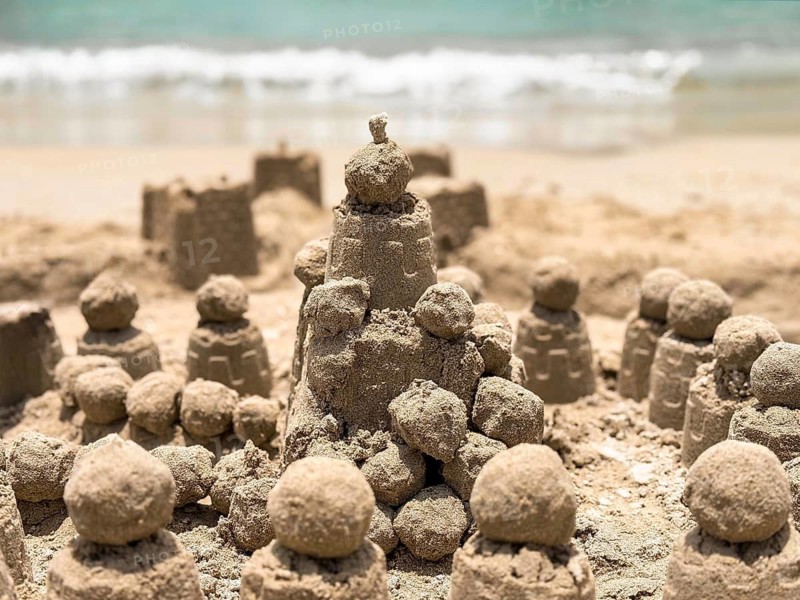 A picturesque scene of detailed sand castles crafted by the seaside, with waves gently lapping in the background under the bright sun, creating a tranquil summer vibe
