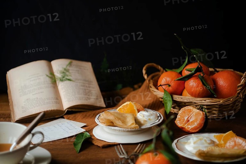 A cozy setting showcases citric crepes accompanied by creamy yogurt, with a basket of fresh oranges and green leaves adding a vibrant touch. A vintage book lies open, enhancing the rustic charm of the dark wooden table