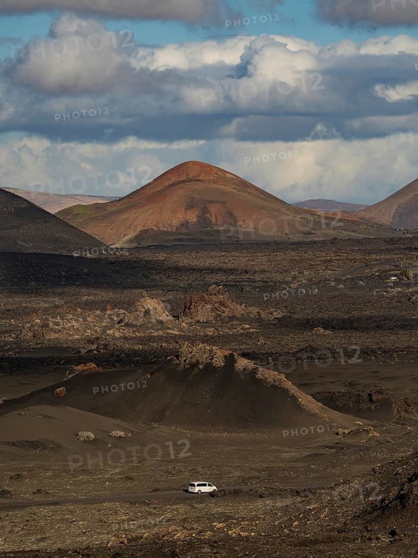 Dramatic volcanic landscape and vast skies of Timanfaya National Park in Lanzarote, showcasing rugged terrain under a dynamic sky with a distant solitary vehicle navigating through