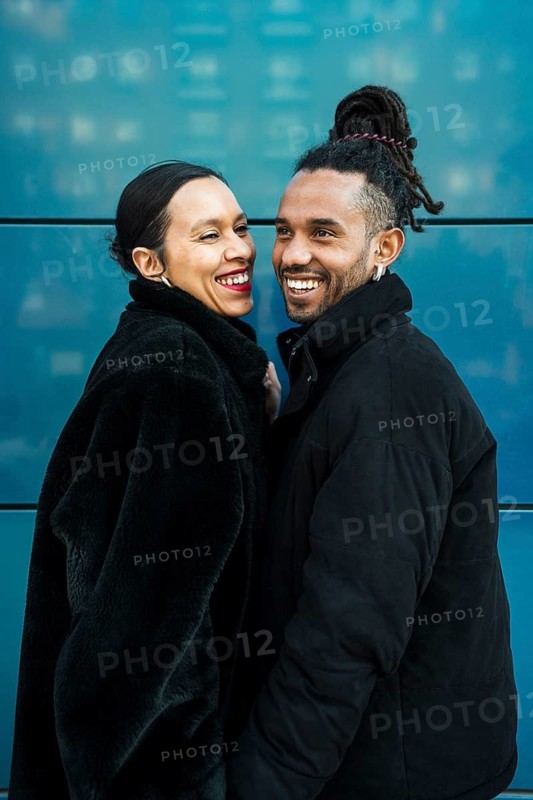 A joyful multicultural couple dressed in stylish winter coats enjoys a happy moment together, set against a modern blue backdrop, highlighting love and diversity