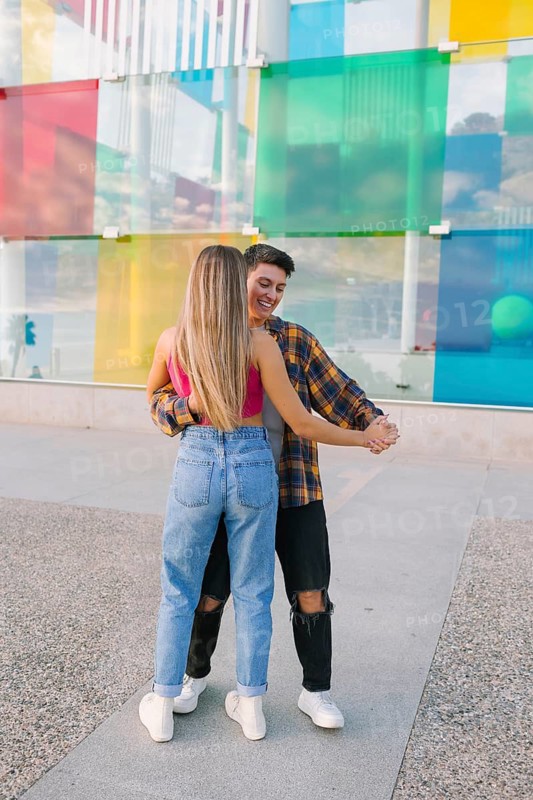 A joyful lesbian couple dances together in an urban setting, smiling and holding hands. The vibrant glass wall behind them adds a dynamic and artistic backdrop, symbolizing love and freedom
