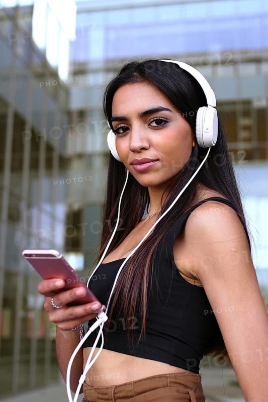 A young woman stands outdoors in an urban setting, wearing headphones and holding her smartphone. She is dressed in a casual black top, appearing relaxed and content