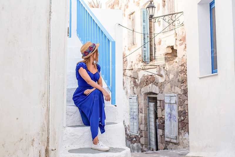 Woman in a vibrant blue dress and straw hat sits on white stairs, admiring the charming architecture of a Greek village in Plaka, Milos island. The scene exudes calm and elegance
